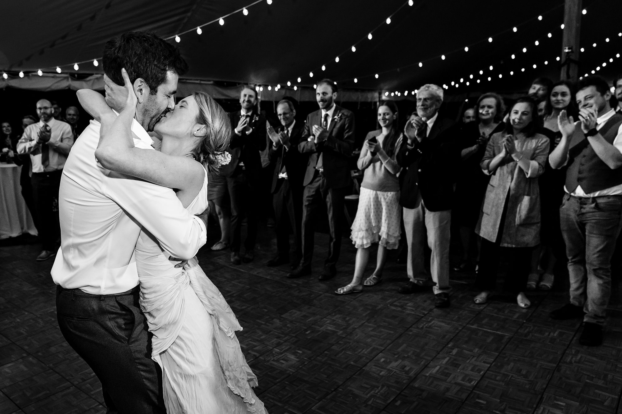 A wedding couple kiss on the dance floor while their family and friends clap.