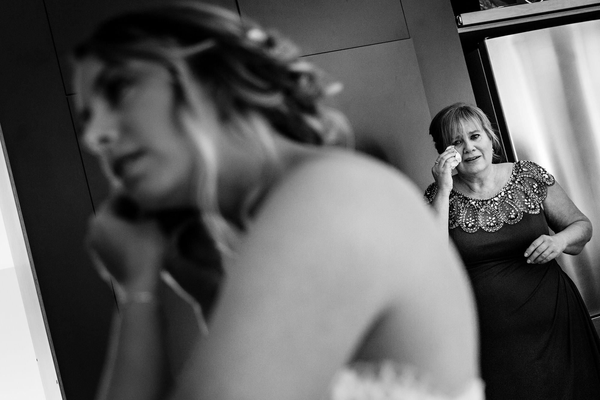 The mother of the bride cries while the bride finishes getting dressed on her wedding day.