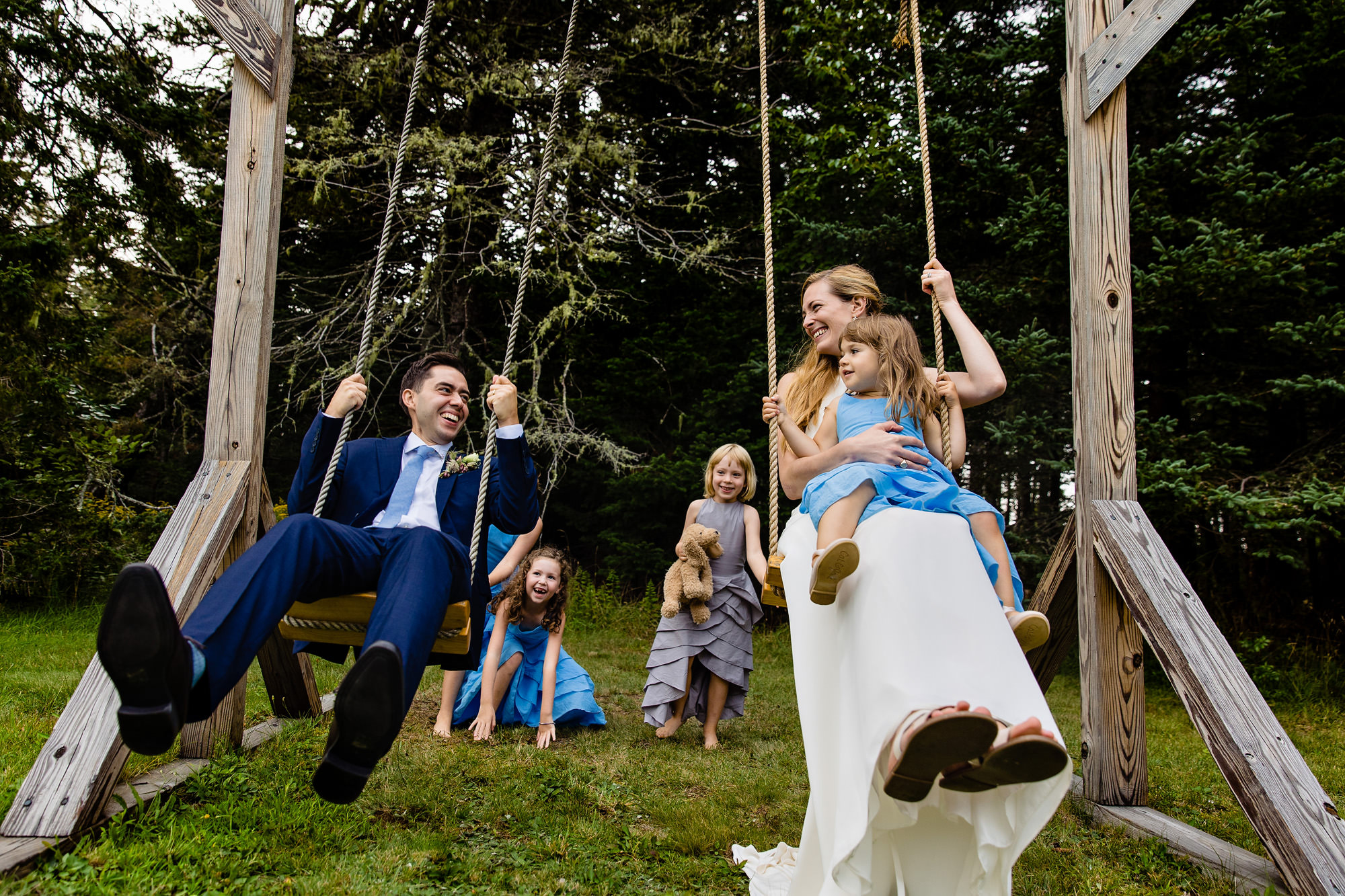 A wedding couple swing on a swing set while their flower girls push them.