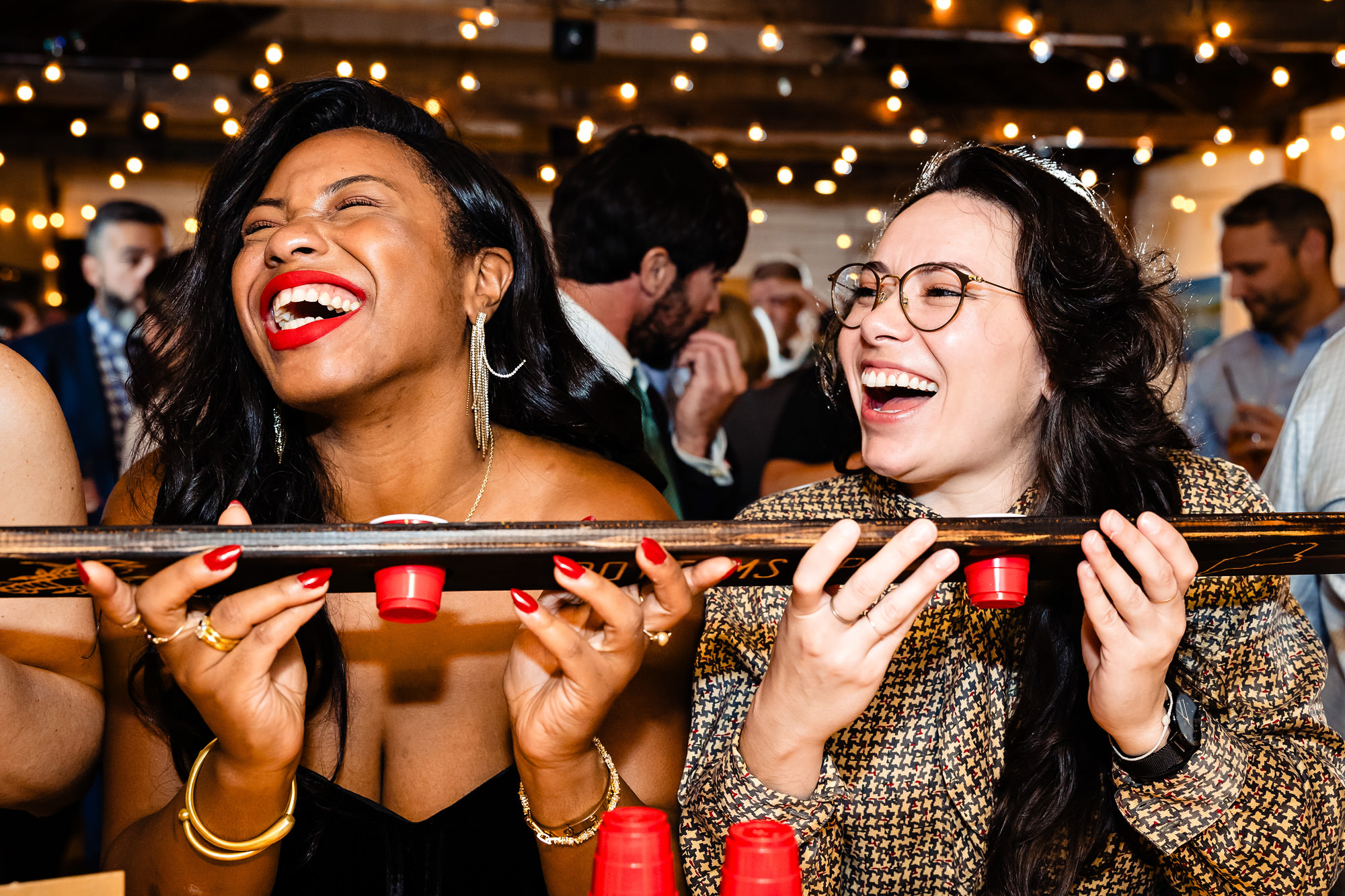 Two women hold a shotski at a wedding while laughing.