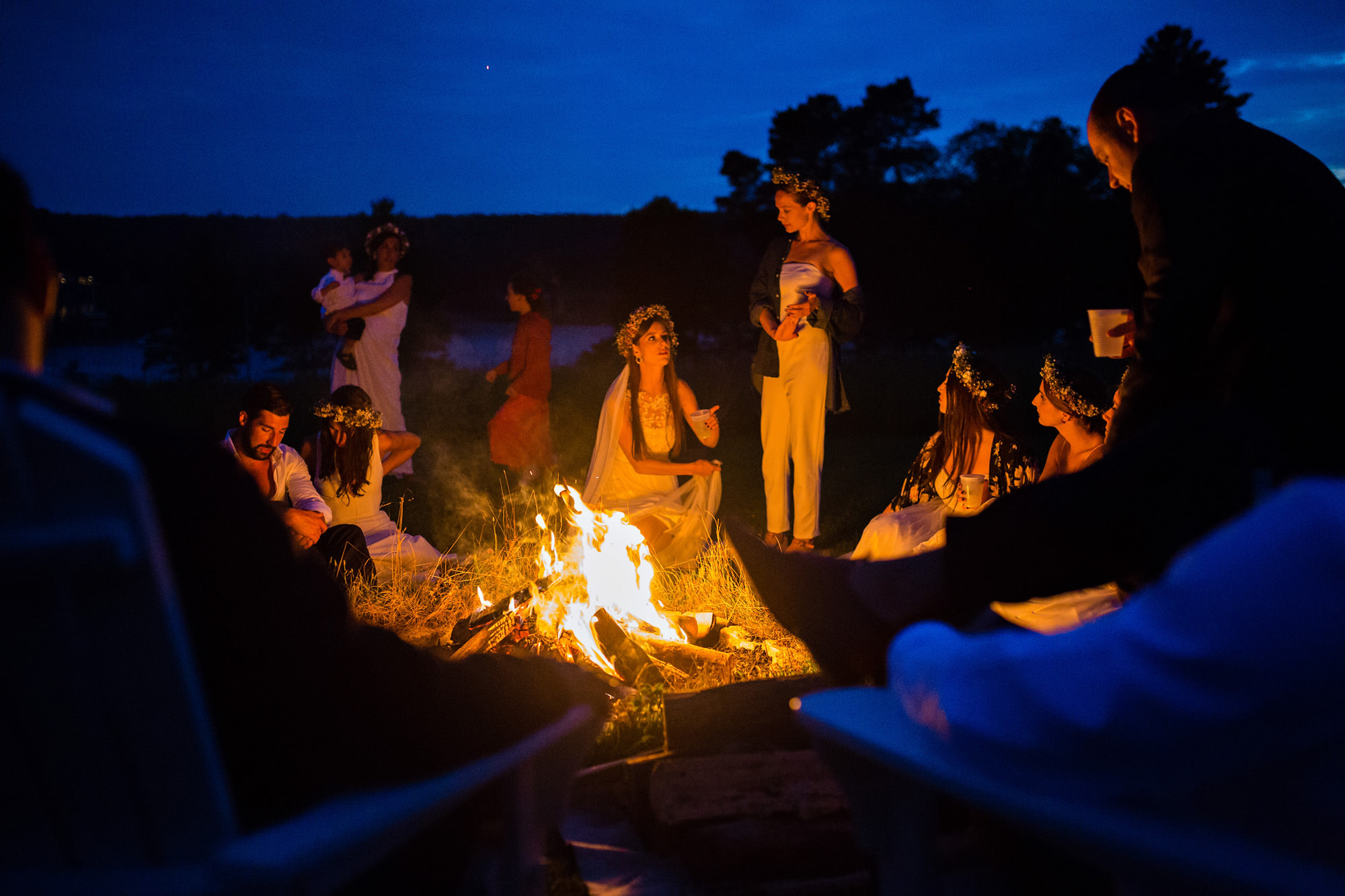 A wedding couple sits by a fire at twilight with their wedding party.